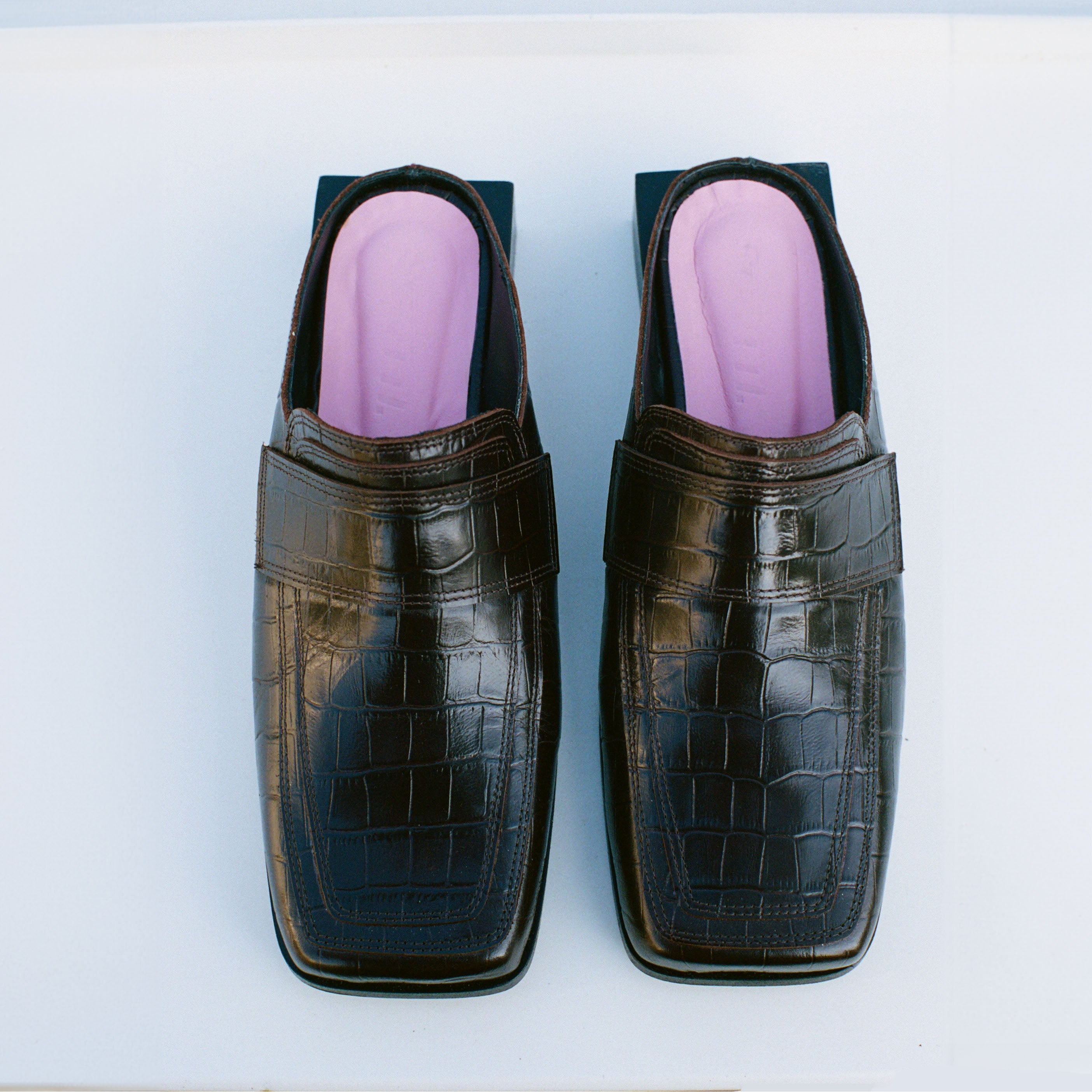 A pair of SCOTTI dark brown, crocodile-patterned leather loafers with square toes sits on a white surface. They feature striking pink insoles visible from the top view.