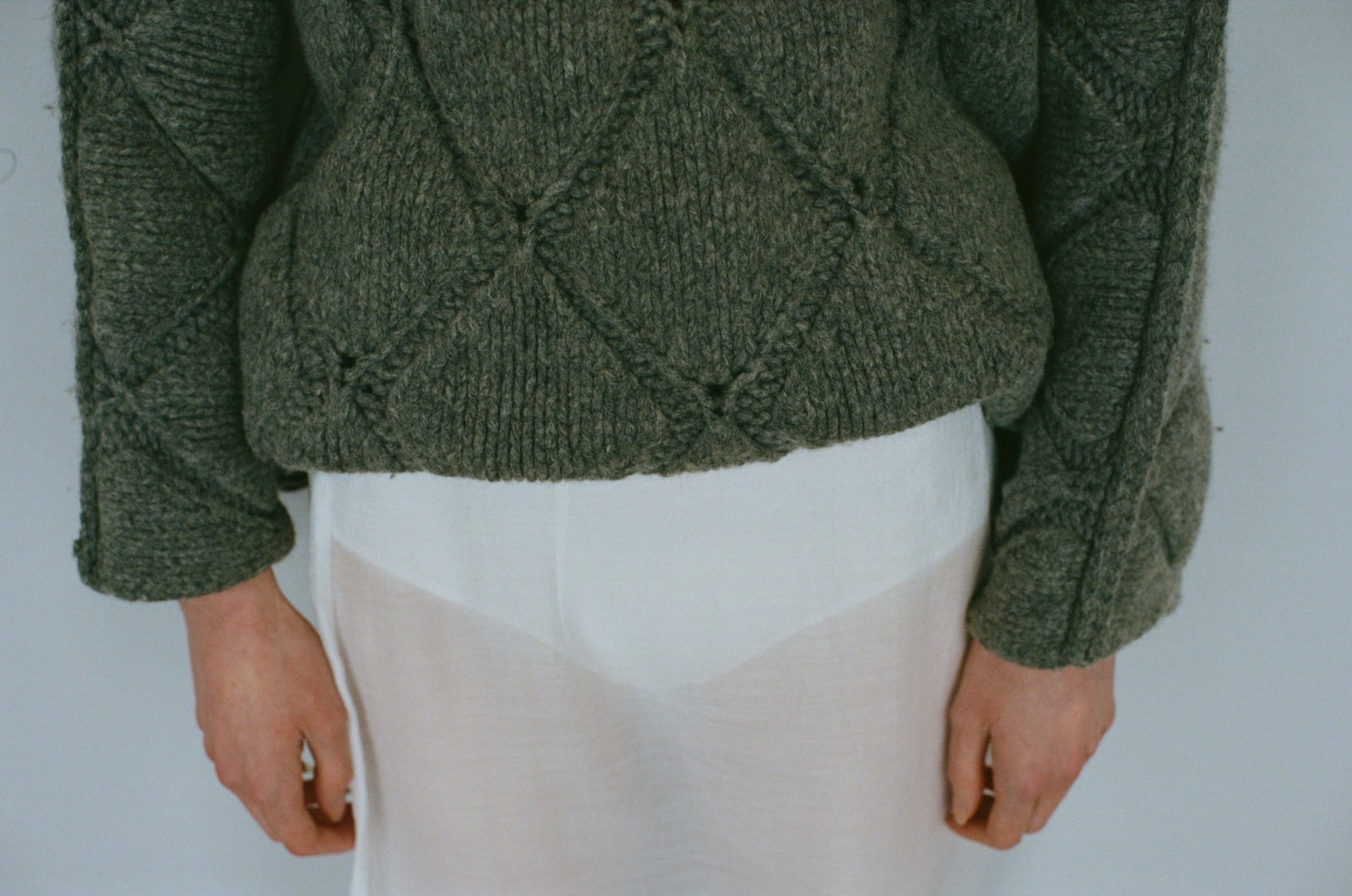 close-up of persons torso wearing a grey thick knitted sweater and a sheer white skirt and het hands.