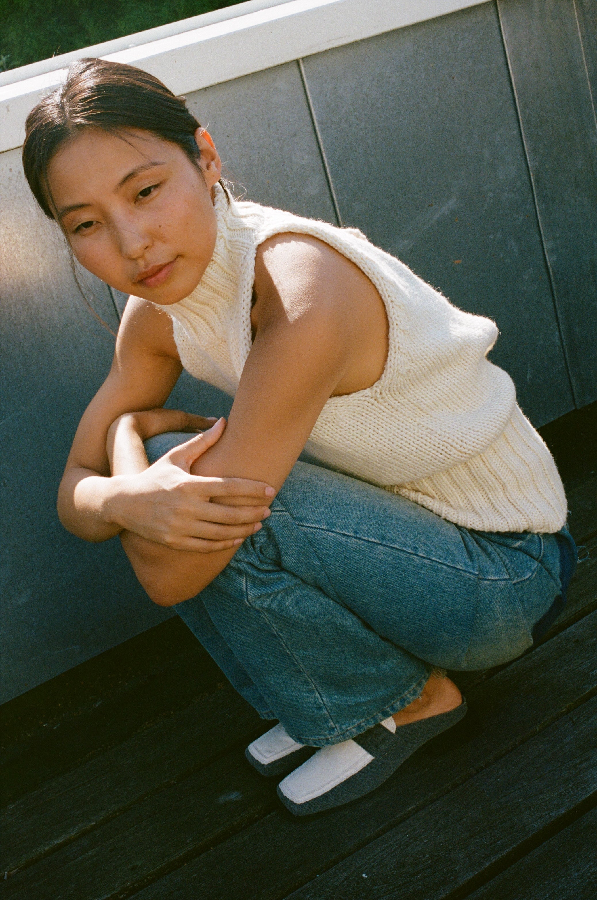 Woman crouching outdoors wearing SCOTTI square toe loafer mules in grey and white, styled with blue jeans and a sleeveless cream knit top.