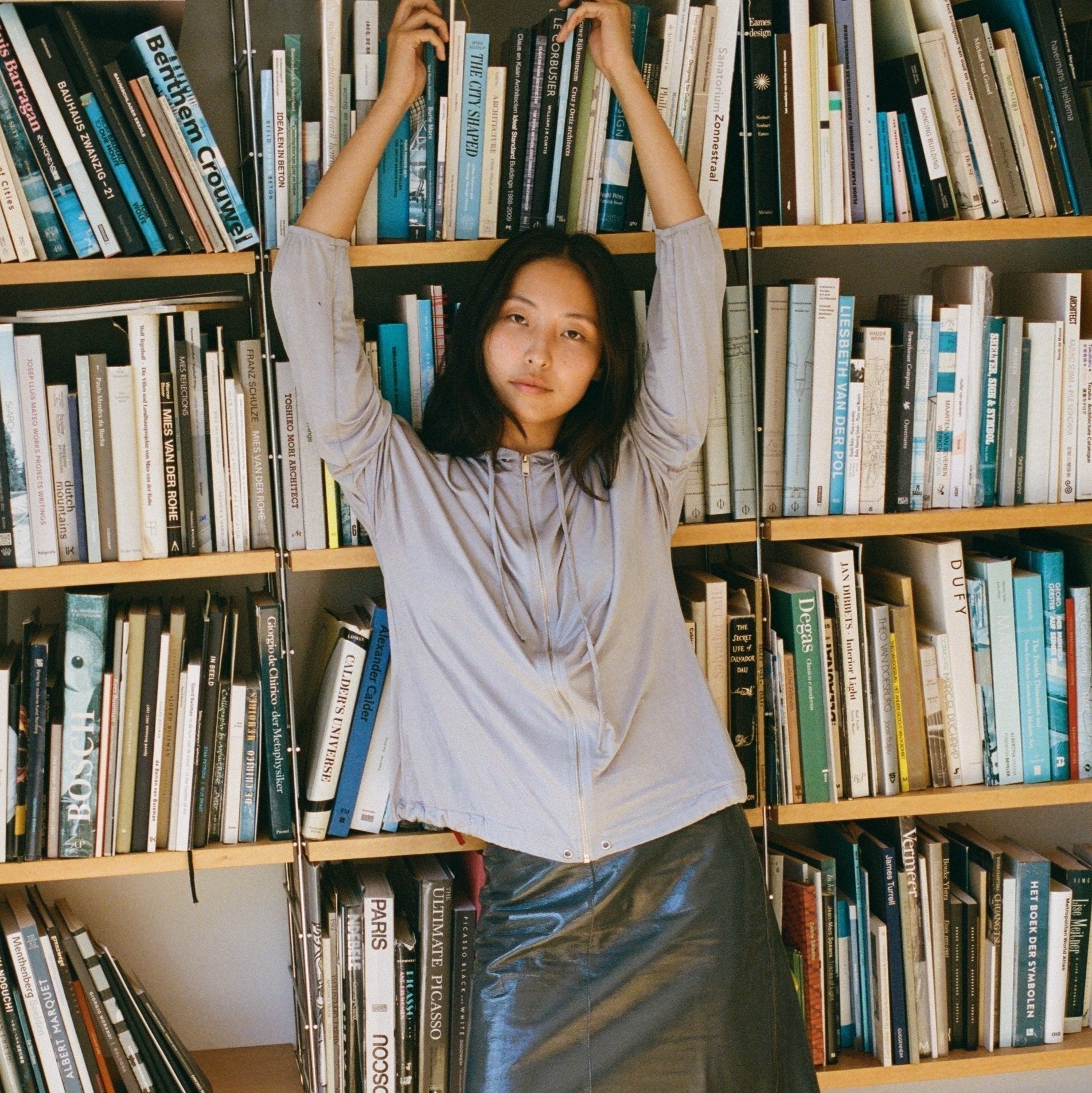 Women with long brown hair leans back with her arm above her head on a mid century design bookshelf with chrome and wood, filled with books. She wears a light grey vest and a shiny grey leather skirt