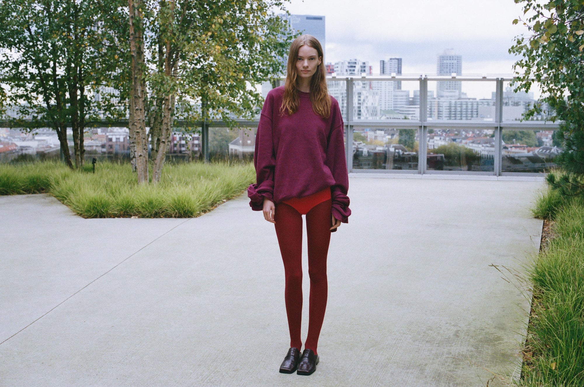 A person stands outdoors on a concrete rooftop surrounded by grass and trees, wearing a burgundy sweater, red leggings, and dark brown loafers from SCOTTI.  In the background, the city skyline with its towering buildings looms under a cloudy sky.