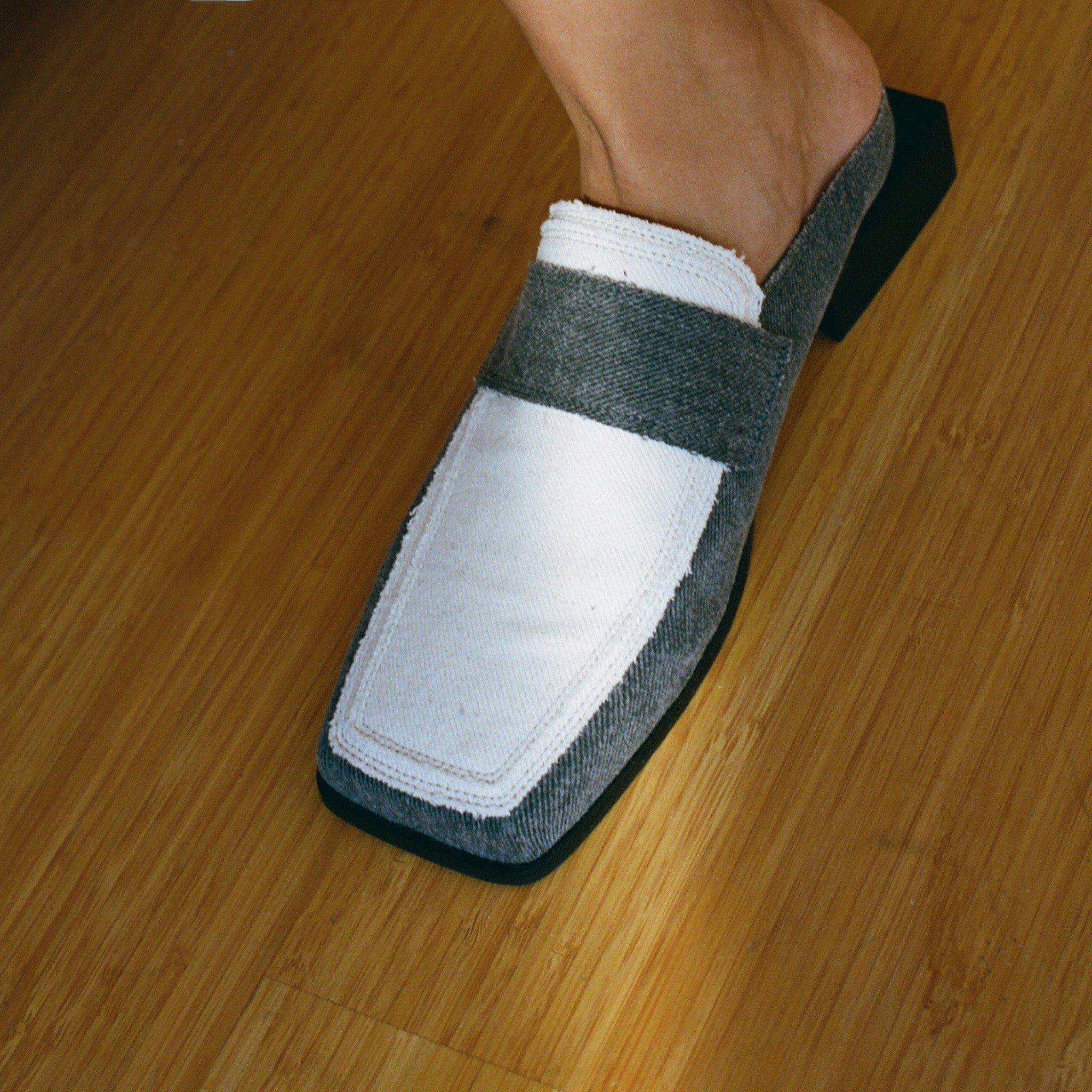 A close-up of a persons foot wearing a SCOTTI loafer with a chic square toe. This stylish shoe is made of a white denim upper and grey denim sides, contrasted by a black sole and square heel, resting on a wooden floor.