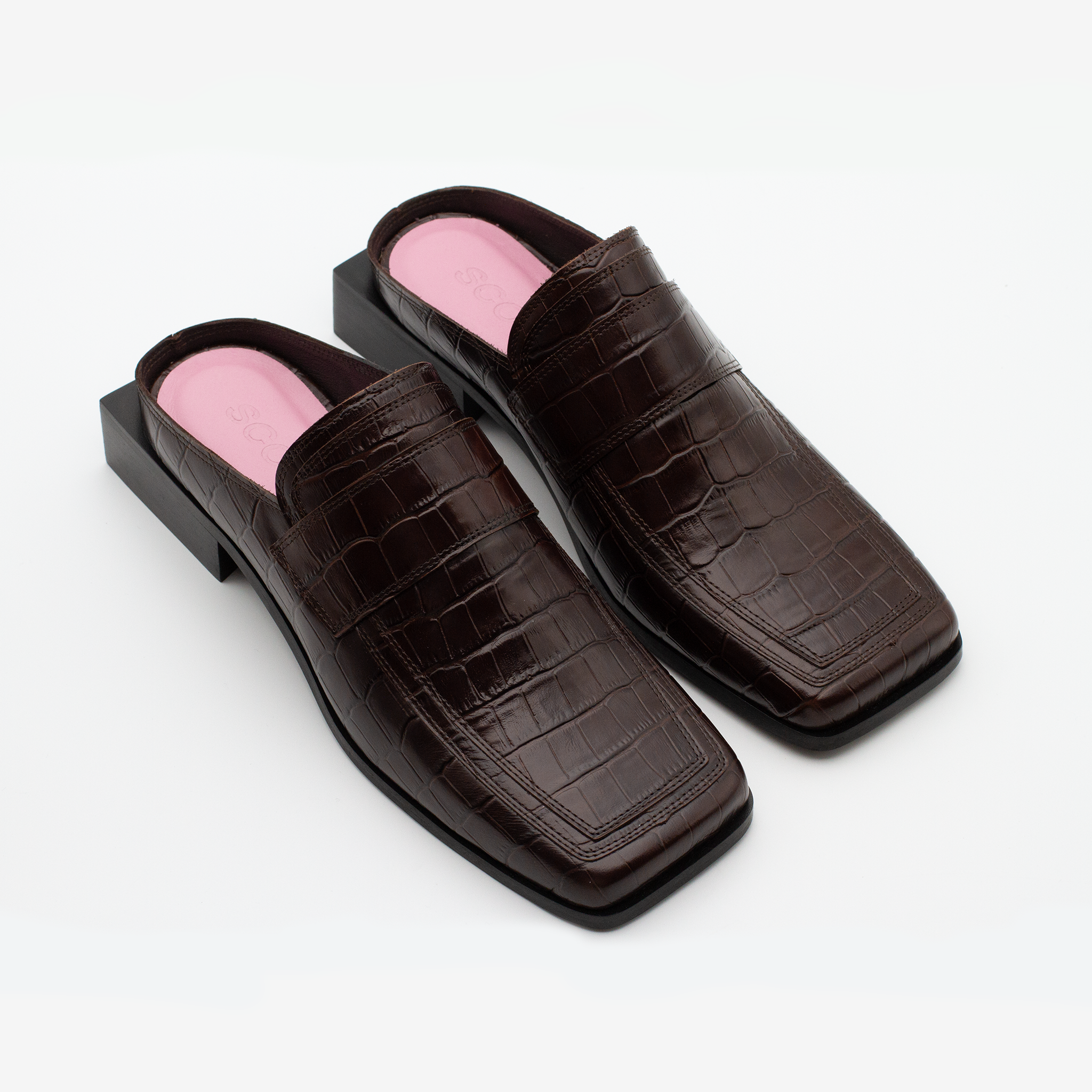 A pair of SCOTTI dark brown, crocodile-patterned leather slip-on loafers with square toes rest on a white background. The shoes feature black square block heel and a pink insole. 