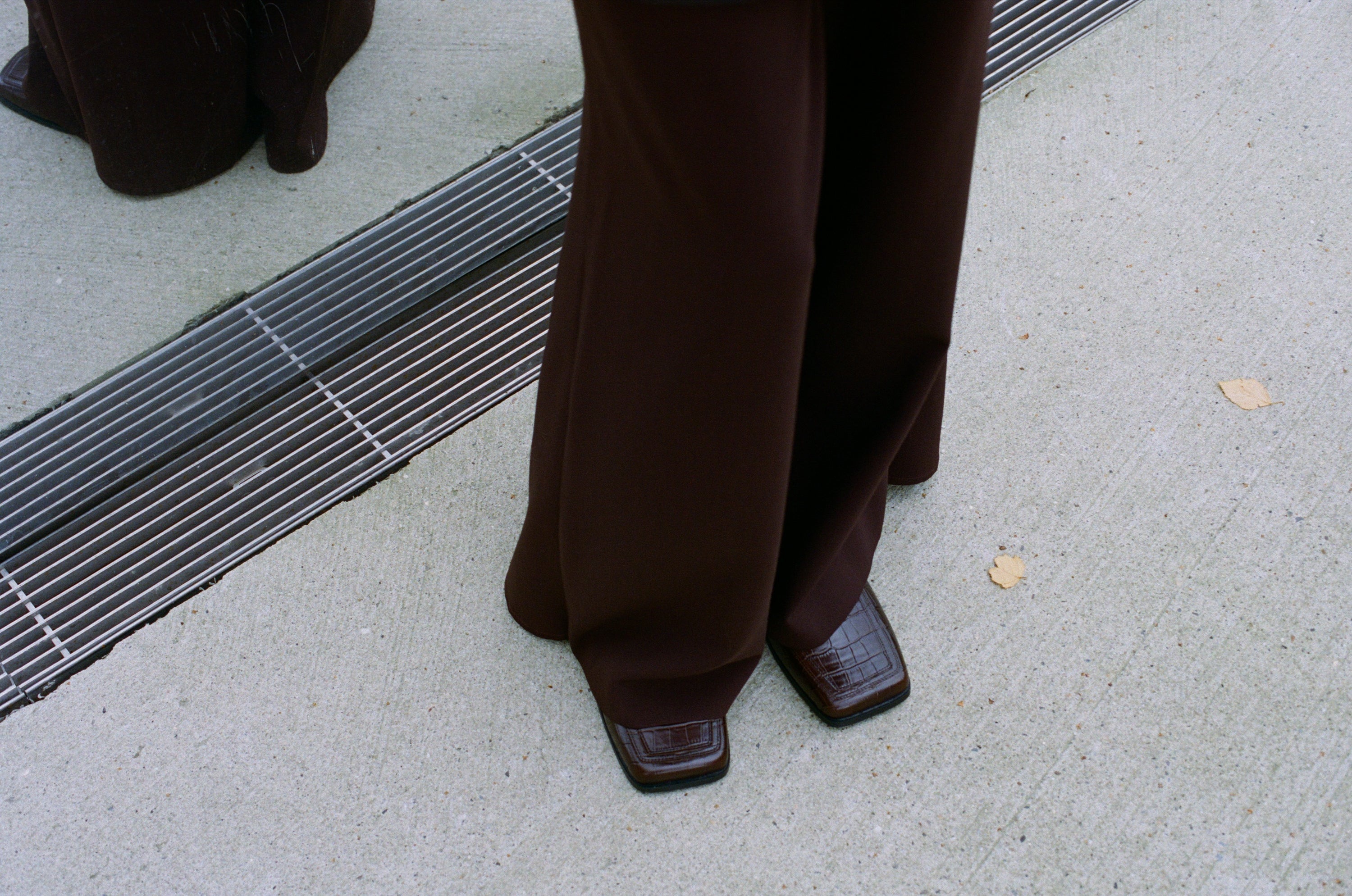 A person wearing wide-legged brown pants and square toe loafer mules from SCOTTI in brown croco embossed leather, stands on a concrete roof of Depot Boijmans van Beuningen near a metal grate, with a few small leaves scattered nearby.